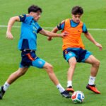 Dos futbolistas disputan el balón en un entrenamiento, uno de ellos con peto naranja.