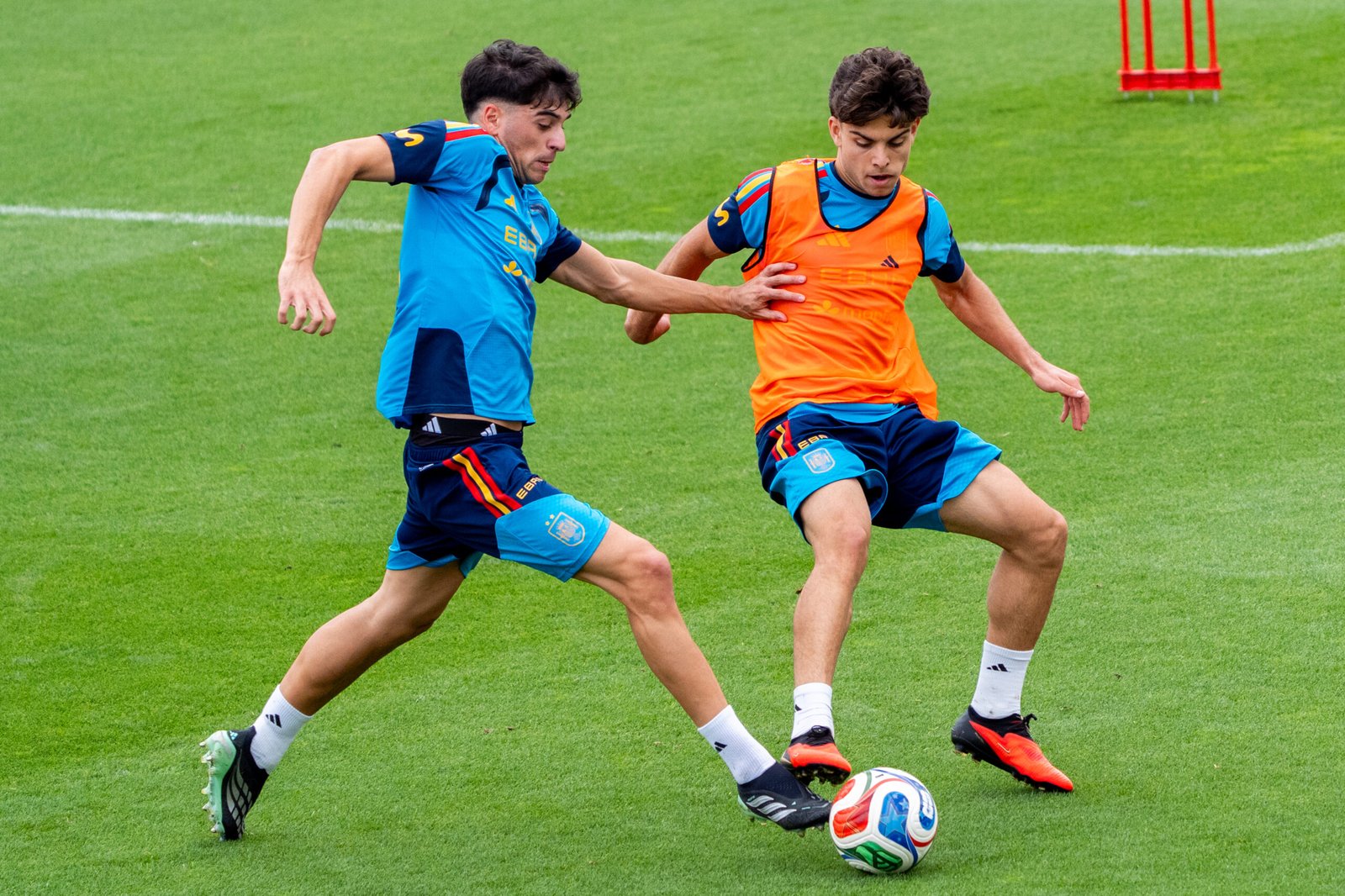 Dos futbolistas disputan el balón en un entrenamiento, uno de ellos con peto naranja.