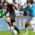 Dos futbolistas disputan el balón sobre el césped; uno viste de negro y rojo y el otro de celeste.