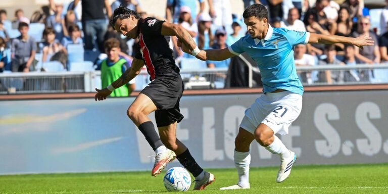 Dos futbolistas disputan el balón sobre el césped; uno viste de negro y rojo y el otro de celeste.