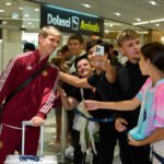 Un futbolista de España posa con aficionados junto al cartel de llegadas de un aeropuerto