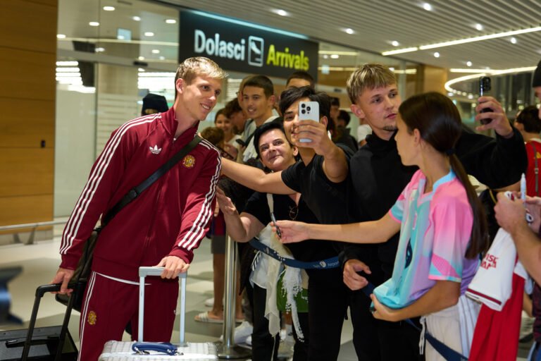 Un futbolista de España posa con aficionados junto al cartel de llegadas de un aeropuerto